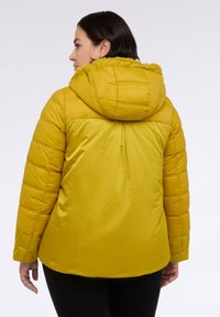 Žlutá péřová bunda s kapucí lemovanou kožešinou, s prošívanou strukturou a hladkým povrchem, nošená přes černé kalhoty.