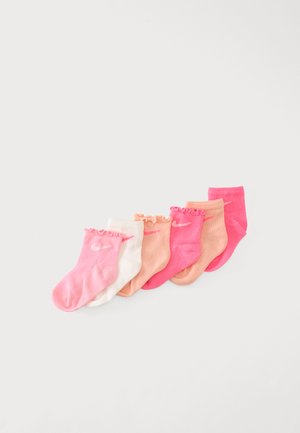 Six paires de chaussettes pour bébé dans des tons de rose, pêche et blanc avec des logos Nike, disposées en diagonale sur un fond blanc uni.