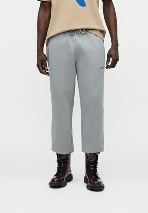 Muž nosiaci svetlosivé skrátené tepláky so šnúrkou a tmavohnedé šnurovacie topánky na jednoduchom bielom pozadí.