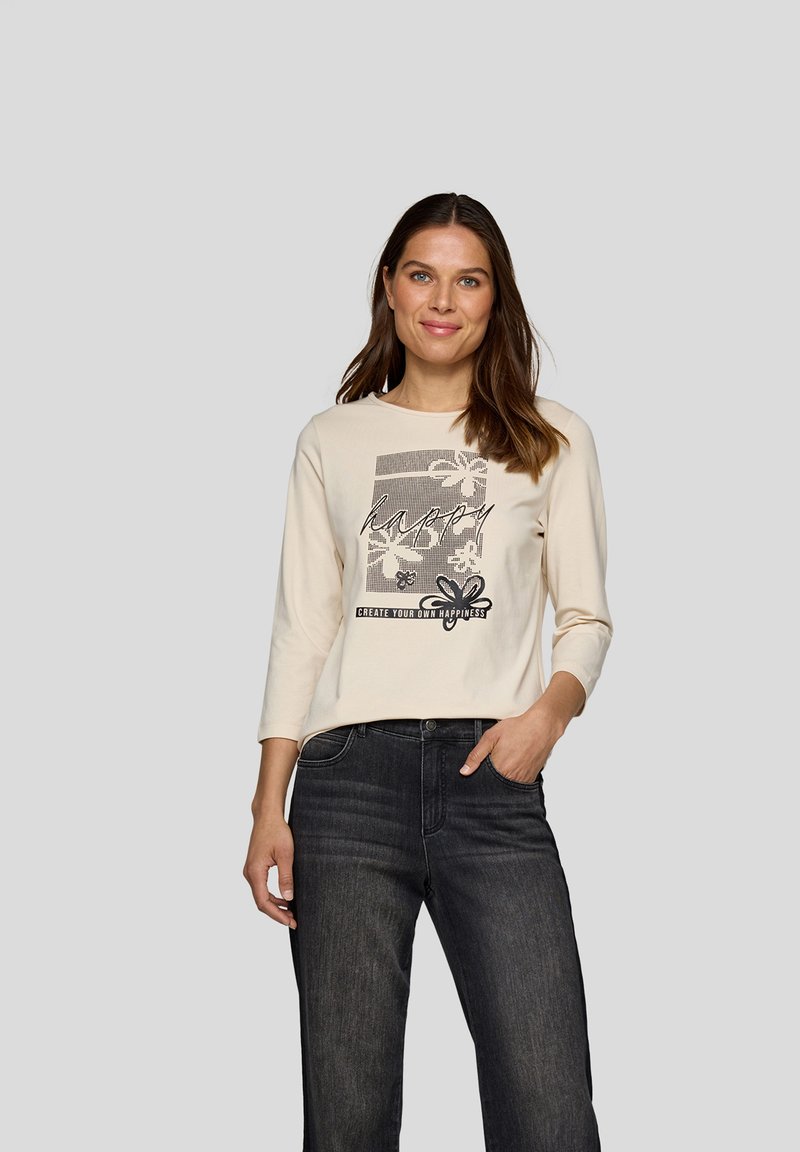 Langärmliges cremefarbenes Oberteil mit einem grafischen Druck und floralen Akzenten sowie Text. Kombiniert mit dunklen Jeans für einen lässigen Look.