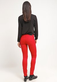 Czerwone wąskie dżinsy wykonane z denimu, o gładkiej teksturze. W połączeniu z czarną bluzką z długim rękawem i czarnymi butami, widok z tyłu.
