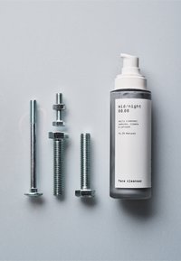 Gezichtsreiniger in een matgrijze fles met een pomp, vergezeld door vier metalen schroeven en bouten op een lichte ondergrond.