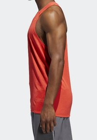 Rotes sportliches Tanktop mit gerippter Textur, breiten Armausschnitten und lockerer Passform, kombiniert mit grauen Shorts. Seitliche Ansicht dargestellt.