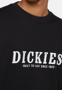 Černá mikina s žebrovaným výstřihem a bílým potiskem. Text uvádí "DICKIES - BUILT TO LAST SINCE 1922." Tkanina vypadá hladce.