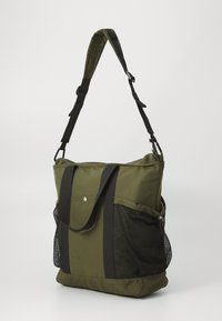 Sac fourre-tout vert olive en tissu avec des sangles noires réglables, un rabat à l'avant et une poche en maille sur le côté. Design simple et fonctionnel.