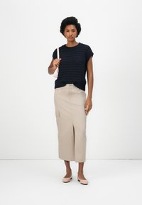 Navy gestricktes kurzärmliges Oberteil, beige mittellange Rock mit vorderem Schlitz und Taschen und hellrosa Ballerinas. Das Model hält eine kleine rosa Tasche.