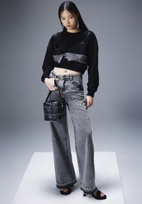 Sudadera negra recortada con superposición de denim, pantalones de mezclilla anchos grises y un bolso tipo cubo negro. Modelo lleva sandalias de tacón negras.