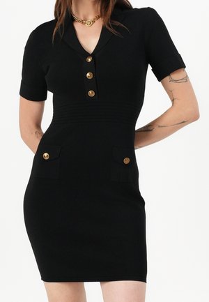 Robe noire ajustée à manches courtes, boutons dorés à l'avant et poches, portée avec un collier chaîne doré.