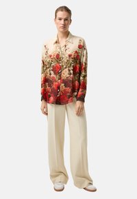 Seidenbluse mit floralem Muster in Rot und Creme, mit durchgehender Knopfleiste und langen Ärmeln. Kombiniert mit cremefarbenen Hosen mit weitem Bein.