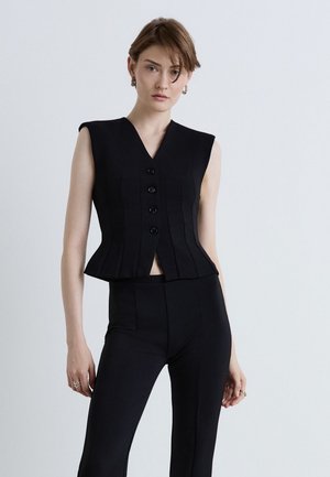 Jeune femme aux cheveux courts portant un haut noir sans manches boutonné et un pantalon noir assorti sur un fond clair uni.