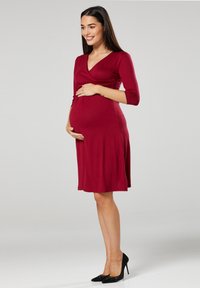 Schwangerschaftsabendkleid in Burgunderrot mit Wickeldesign, Dreiviertelärmeln und schwingendem Rock. Getragen mit schwarzen High Heels.
