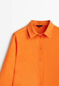Chemise orange à manches longues avec un col boutonné, présentant un tissu texturé et des boutons assortis le long de la patte de boutonnage.