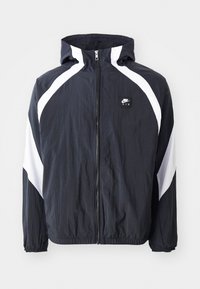 Musta tuulitakki, jossa valkoiset yksityiskohdat. Ominaisuuksiin kuuluu vetoketjullinen etuosa, huppu ja joustavat hihansuut. Valmistettu kevyestä, kuvioidusta kankaasta.