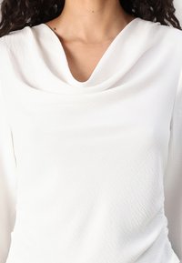 Haut blanc avec décolleté drapé et manches longues, confectionné en tissu texturé présentant des vagues subtiles, créant un tombé doux.