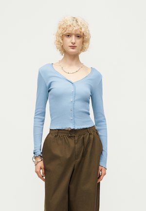 Osoba s kučeravými blond vlasmi má oblečený svetlomodrý zapínaný top s dlhým rukávom a výstrihom do V, hnedé nohavice s vysokým pásom, strieborný náhrdelník a náramky.