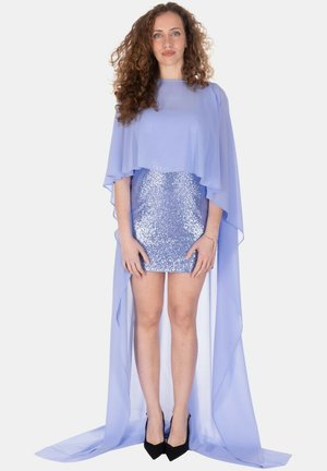 Donna con capelli ricci che indossa un mini abito blu lucido con paillettes, un mantello trasparente e fluido e tacchi alti neri, in piedi su sfondo bianco.