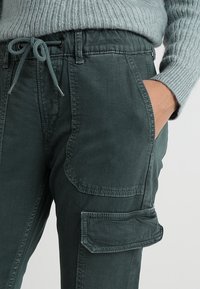 Primer plano de una persona que lleva pantalones cargo verde oscuro con un gran bolsillo lateral y un suéter de punto gris claro, con la mano en el bolsillo.