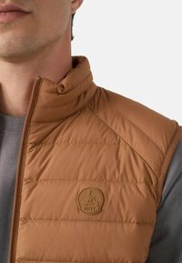 Gilet imbottito marrone con colletto alto, texture trapuntata e logo JOTT. Indossato sopra una maglia grigia, con un design aderente.