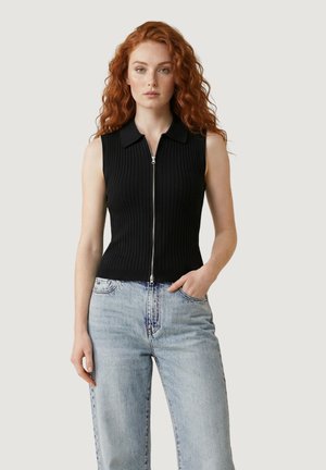 Donna dai capelli rossi che indossa un top nero senza maniche con zip frontale e jeans aderenti a vita alta azzurri, in piedi davanti a uno sfondo semplice.