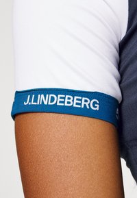 Kortärmad skjorta med vit kropp och mörkgrå detaljer, utrustad med en blå manschett med vit "J.LINDEBERG"-logotyp. Mjuk textur.