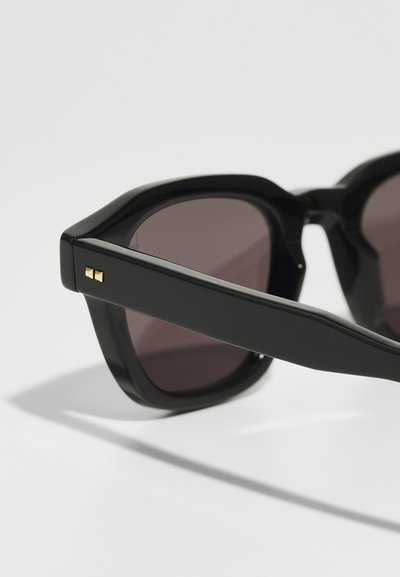 Black sunglasses with a chunky design, featuring dark lenses and gold accent on the temples. Smooth plastic texture with angular shapes.