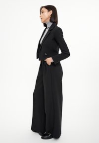 Blazer negro corto con bolsillos laterales, combinado con pantalones negros de pierna ancha. Suéter de cuello alto gris debajo, zapatos negros brillantes. Tejido suave.