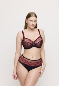 Kobieta ubrana w czarny i czerwony koronkowy biustonosz oraz pasujące majtki w wysokim stanem, stojąca na tle jednolitego, neutralnego koloru.