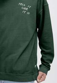 Sudadera verde de manga larga con texto blanco en el pecho que dice "DÍLO COMO ES". Fabricada con un material suave y con un corte relajado.