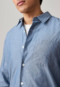 Camicia a righe blu con bottoni, realizzata in tessuto leggero, con tasca, colletto e bottoni bianchi, caratterizzata da strisce verticali di diverse tonalità.