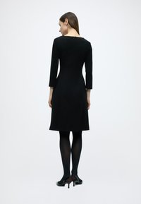Μαύρο φόρεμα σε γραμμή Α με μανίκια τριών τετάρτων και φερμουάρ στο πίσω μέρος. Το ύφασμα φαίνεται λείο, σε συνδυασμό με διαφανείς μαύρες καλσόν και παπούτσια με τακούνι.