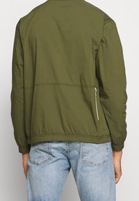 Osoba oblečená v olivově zelené bundě s kapsou na zip na pravé zadní straně a v světle modrých džínách.