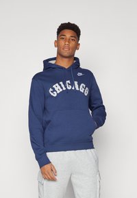 Mørkeblå hettegenser laget av bomull, med "CHICAGO" i hvite bokstaver, frontlomme og snorhette. Kombinert med grå joggebukser.