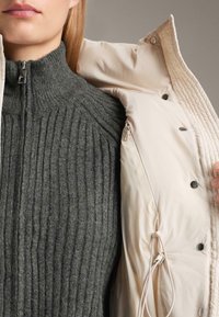 Osoba nosi tmavosivý rebrovaný sveter na zips a drží otvorenú svetlobéžovú bundu so zatrhávacími gombíkmi a vnútorným vreckom na zips.