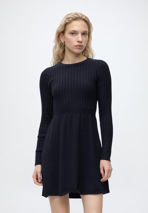 Jeune femme aux cheveux blonds porte une robe noire en maille ajustée à manches longues avec une jupe évasée sur un fond blanc uni.