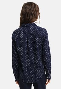 Navy button-up shirt with white polka dots, featuring a classic collar, long sleeves, and a curved hem. Smooth fabric texture.