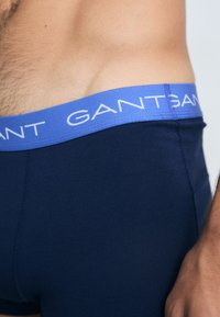 Tamno plave uske bokserice od glatkog materijala, s mekim, elastičnim svjetloplavim pojasom s bijelim "GANT" brendiranjem.