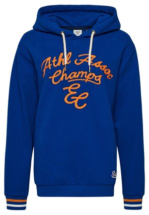 Blauer Sweatshirt mit gesticktem Text "Superdry Assoc" und geripptem Bündchen mit weißen, orangefarbenen und blauen Streifen, getragen mit dunkler Jeans.