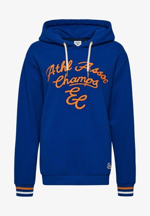 Blauwe trui met geborduurde tekst "Superdry Assoc" en geribbelde manchetten met witte, oranje en blauwe strepen, gedragen met donkere jeans.