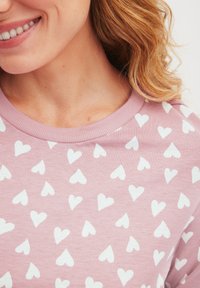 Hellrosa Sweatshirt mit einem weißen Herzmuster. Der Stoff ist weich, mit einem runden Ausschnitt und geripptem Kragen.