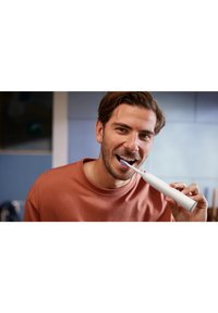 Brosse à dents électrique avec un manche blanc et des poils bleus tenue par une personne portant une chemise couleur rouille, souriante tout en se brossant les dents.