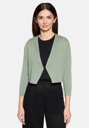 Cardigan vert clair en tissu doux, avec une coupe courte et des manches trois-quarts, porté par-dessus un haut noir.