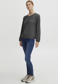 Grå langermet bluse med rynkede detaljer, kombinert med tettsittende blå jeans og beige trening sneakers med lyse detaljer.