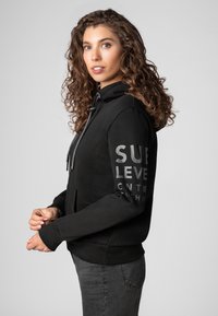 Zwarte hoodie met een aantrekkoord in de capuchon, een voork pocket en zilveren opdruk op de mouw. Gemaakt van zachte stof met ribgebreide mouwen en onderrand.