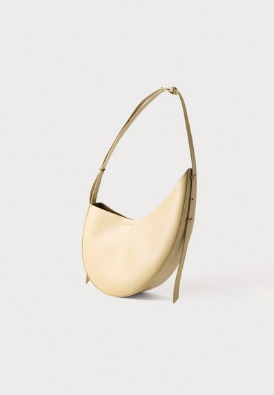 Μπεζ δερμάτινη τσάντα ώμου σε σχήμα ημισελήνου με ρυθμιζόμενο ιμάντα, εμφανίζεται όρθια σε λευκό φόντο.