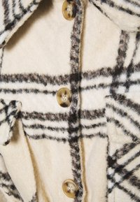 Krémový vlněný kabát s černobronzovým kostkovaným vzorem, se třemi knoflíky z želvoviny a kapsou s klopou.