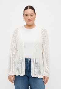 White knitted cardigan with an open front, featuring a lace-like texture and scalloped hem. Worn over a plain white shirt and blue jeans.