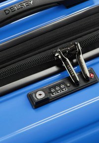 Valise rigide bleue avec deux tirettes de fermeture à glissière, un verrou à combinaison et une fermeture à glissière texturée noire. La marque "Delsey" est visible.