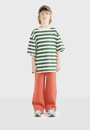 T-shirt rayé vert et blanc, coupe oversize, associé à un pantalon large orange avec couture sur les côtés. Une casquette grise et des baskets grises complètent le look.