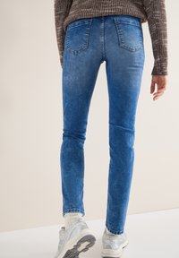 Personne portant un jean bleu slim, des chaussettes blanches, des baskets argentées et un pull en maille marron, debout devant un fond beige uni.