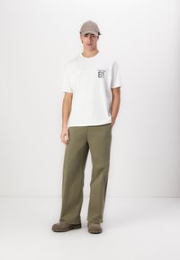 Hvit t-skjorte med korte ermer og trykt design, sammen med olivengrønne vide bukser og beige sneakers. Grå caps fullfører antrekket.